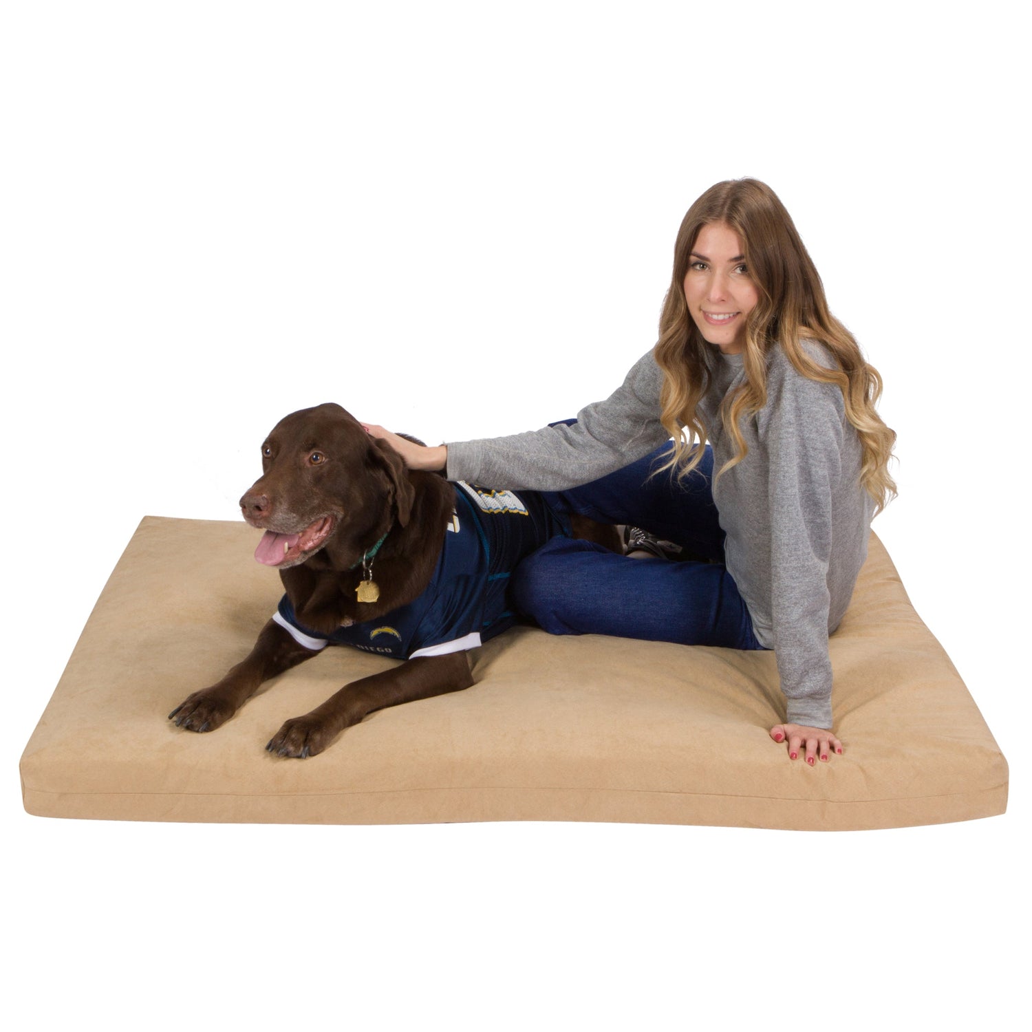 A woman with long brown hair in a gray sweater smiles and pets a large brown dog wearing a navy blue jersey as he lies on the Orthopedic Memory Foam Dog Bed, looking at the camera.