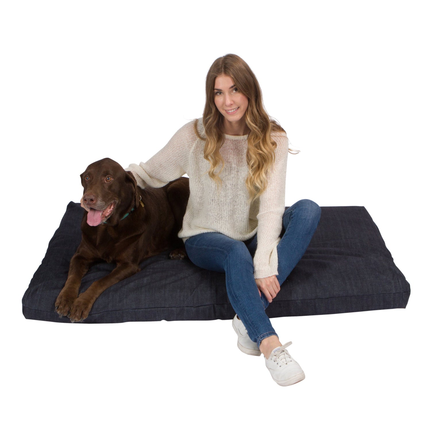 A woman with long, wavy hair sits beside a brown dog on an Orthopedic Memory Foam Dog Bed, smiling as she rests her hand on the dogs back. Both look ahead; the dogs tongue is out.